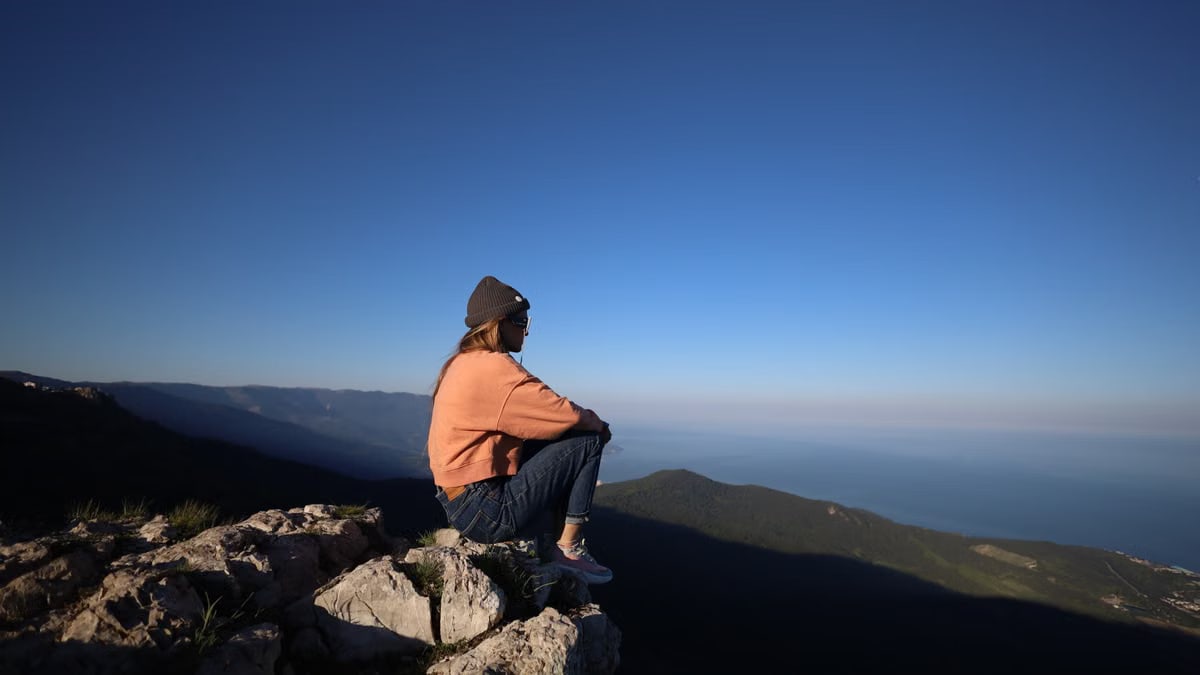 A woman sits on a mountain cliff, enjoying a panoramic view under a clear blue sky.